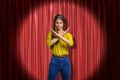 Young determined woman wearing jeans and yellow shirt making rejection gesture on red stage curtains background