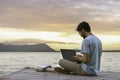 Young digital nomad man sitting on wooden pier at sea working on internet remotely at sunset - Traveling with a computer - Online Royalty Free Stock Photo