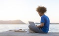 Young digital nomad man sitting on wooden pier at sea working on internet remotely at sunset - Traveling with a computer - Online Royalty Free Stock Photo