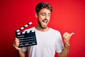 Young director man with beard making movie using clapboard over isolated red background pointing and showing with thumb up to the