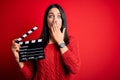 Young director woman with blue eyes making movie holding clapboard over red background cover mouth with hand shocked with shame