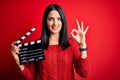 Young director woman with blue eyes making movie holding clapboard over red background doing ok sign with fingers, excellent