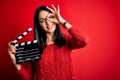 Young director woman with blue eyes making movie holding clapboard over red background with happy face smiling doing ok sign with