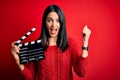 Young director woman with blue eyes making movie holding clapboard over red background screaming proud and celebrating victory and