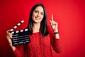 Young director woman with blue eyes making movie holding clapboard over red background surprised with an idea or question pointing