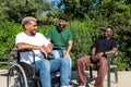 Young disabled African American man in a wheelchair and his male friends talking sitting on a bench in city park. Royalty Free Stock Photo