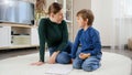 Young displeases mother shouting and talking strictly to her son doing homework on carpet at living room. Concept of