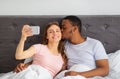 Young diverse couple with cellphone kissing while taking selfie in bed at home Royalty Free Stock Photo