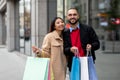 Young diverse couple with paper bags walking near store windows, shopping together, smiling at camera outdoors Royalty Free Stock Photo