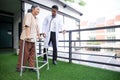 Young doctor assistance elderly patient while guidance therapy with walker in garden at hospital.