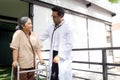 Young doctor assistance elderly patient while guidance therapy with walker in garden at hospital.