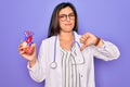 Young doctor cardiology specialist woman holding medical heart over pruple background with angry face, negative sign showing