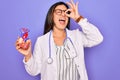 Young doctor cardiology specialist woman holding medical heart over pruple background with happy face smiling doing ok sign with