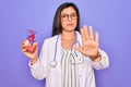 Young doctor cardiology specialist woman holding medical heart over pruple background with open hand doing stop sign with serious