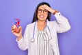 Young doctor cardiology specialist woman holding medical heart over pruple background stressed with hand on head, shocked with