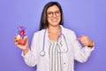 Young doctor cardiology specialist woman holding medical heart over pruple background with surprise face pointing finger to