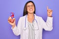 Young doctor cardiology specialist woman holding medical heart over pruple background surprised with an idea or question pointing