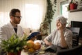 Young doctor consulting and examining senior white-haired woman during home visit