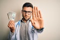 Young doctor man holding a bunch of one dollar banknotes over isolated background with open hand doing stop sign with serious and Royalty Free Stock Photo