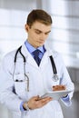 Young doctor man standing at his hospital office, with a stethoscope, using a computer tablet. Perfect medical service Royalty Free Stock Photo