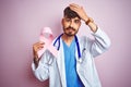Young doctor man with tattoo holding cancer ribbon standing over isolated pink background stressed with hand on head, shocked with Royalty Free Stock Photo