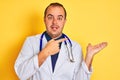 Young doctor man wearing coat and stethoscope standing over isolated yellow background amazed and smiling to the camera while Royalty Free Stock Photo