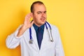 Young doctor man wearing coat and stethoscope standing over isolated yellow background smiling with hand over ear listening an Royalty Free Stock Photo