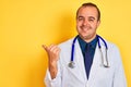 Young doctor man wearing coat and stethoscope standing over isolated yellow background smiling with happy face looking and Royalty Free Stock Photo