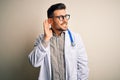 Young doctor man wearing glasses, medical white robe and stethoscope over isolated background smiling with hand over ear listening Royalty Free Stock Photo