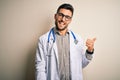 Young doctor man wearing glasses, medical white robe and stethoscope over isolated background smiling with happy face looking and Royalty Free Stock Photo