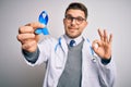 Young doctor man wearing medical coat and holding colon cancer awareness blue ribbon doing ok sign with fingers, excellent symbol Royalty Free Stock Photo