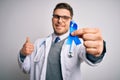 Young doctor man wearing medical coat and holding colon cancer awareness blue ribbon happy with big smile doing ok sign, thumb up Royalty Free Stock Photo