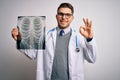 Young doctor man wearing medical coat looking at chest scan radiography over isolated background doing ok sign with fingers,