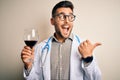 Young doctor man wearing stethoscope drinking a glass of fresh wine over isolated background pointing and showing with thumb up to Royalty Free Stock Photo