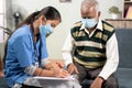 Young doctor or nurse writing prescription during home visiting to sick elder man while both worn face mask due to coronavirus