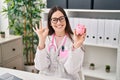 Young doctor woman holding piggy bank doing ok sign with fingers, smiling friendly gesturing excellent symbol Royalty Free Stock Photo