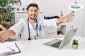 Young doctor working at the clinic using computer laptop looking at the camera smiling with open arms for hug Royalty Free Stock Photo