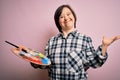 Young down syndrome artist woman holding painter palette and paintbrush over pink background very happy and excited, winner