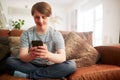 Young Downs Syndrome Man Sitting On Sofa Using Mobile Phone At Home