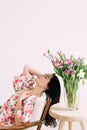 Young dreaming woman sitting in armchair with closed eyes next to vase with flowers bouquet on the table