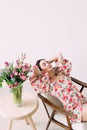 Young dreaming woman sitting in armchair with closed eyes next to vase with flowers bouquet on the table