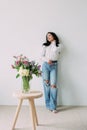 Young dreaming woman standing near wall with closed eyes next to bouquet of flowers on the table