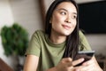 Young dreamy woman looking outside window, holding smartphone and smiling, chatting on phone app Royalty Free Stock Photo