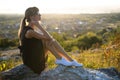 Young elegant woman in black short dress and white sneaker shoes sitting on a rock relaxing outdoors at summer evening. Royalty Free Stock Photo