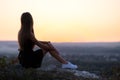 Young elegant woman in black short dress and white sneaker shoes sitting on a rock relaxing outdoors at summer evening Royalty Free Stock Photo