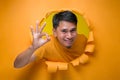 Young entertaining man through torn yellow paper hole showing okay sign with facetious of content and happiness Royalty Free Stock Photo