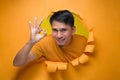 Young entertaining man through torn yellow paper hole showing okay sign with facetious of content and happiness Royalty Free Stock Photo