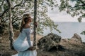 Young European beautiful girl in white bridal marriage dress posing on swing in forest trees on the ocean sea beach Royalty Free Stock Photo