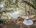 Young European beautiful girl in white bridal marriage dress posing on swing in forest trees on the ocean sea beach Royalty Free Stock Photo