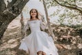 Young European beautiful girl in white bridal marriage dress posing on swing in forest trees on the ocean sea beach Royalty Free Stock Photo